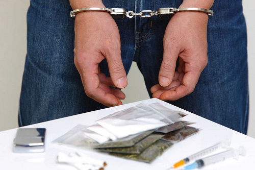 A Man Stands With Hands Cuffed Next to a Table With Drug Products